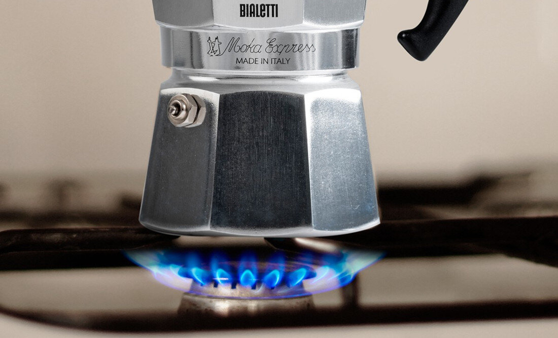 Kawiarka BIALETTI Moka Express znajduje się na palniku gazowym z niebieskim płomieniem. Na kawiarkę widoczny jest napis: \'BIALETTI Moka Express MADE IN ITALY\'.