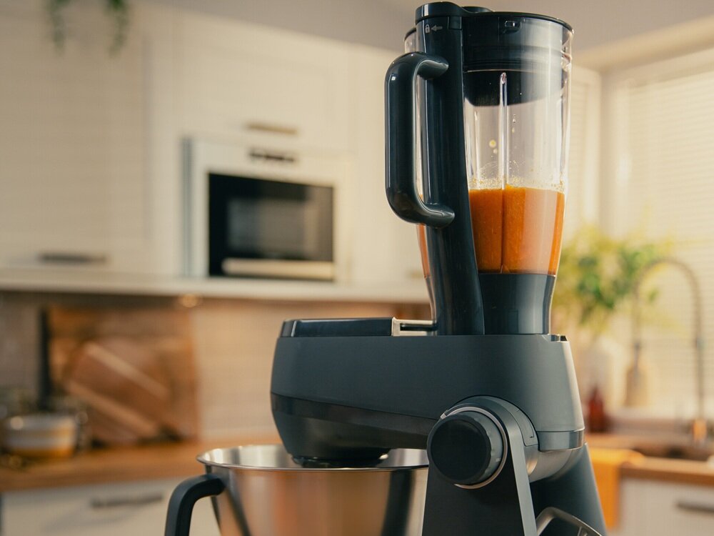 Na obrazie widoczny jest czarny robot kuchenny z blenderem, w którym znajduje się pomarańczowy płyn. W tle znajduje się kuchnia z białymi szafkami i piekarnikiem.
