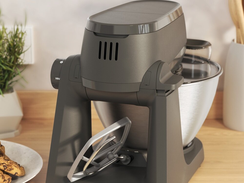 Na blacie kuchennym stoi ciemnoszary robot kuchenny z metalową misą i przystawką do mieszania. W tle widoczne są elementy kuchenne, takie jak doniczka z rośliną i drewniane przybory kuchenne.