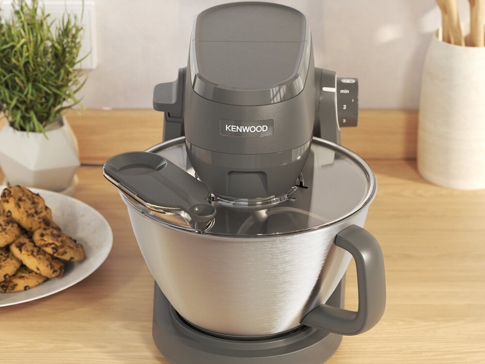Na drewnianym blacie kuchennym stoi robot kuchenny marki Kenwood z dużą metalową misą i pokrywą. Obok znajduje się talerz z ciasteczkami oraz doniczka z rośliną.