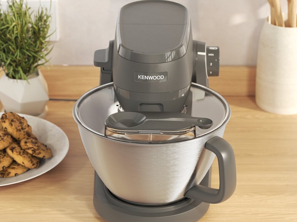 Na drewnianym blacie stoi robot kuchenny marki Kenwood z dużą metalową misą i pokrywą. Obok znajduje się talerz z ciasteczkami oraz doniczka z rośliną.
