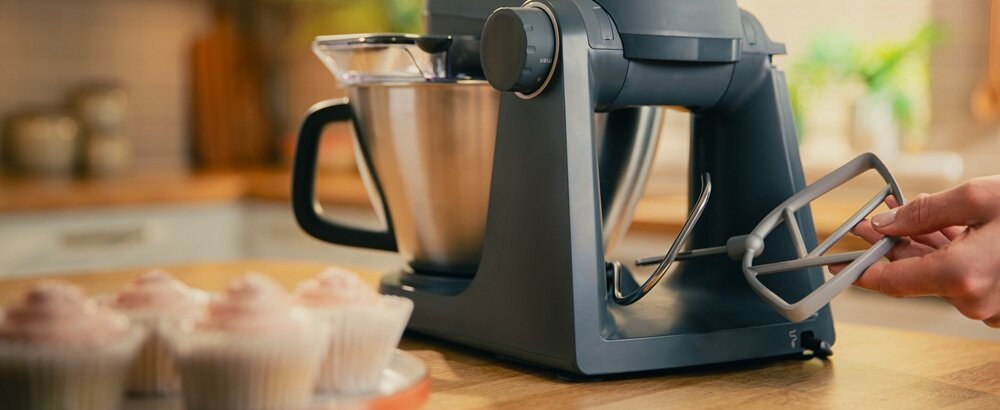 Na blacie kuchennym stoi robot kuchenny z metalową misą i przystawką do mieszania. Obok widoczne są babeczki z różowym kremem.