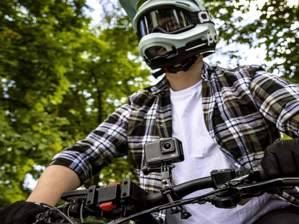 Osoba w kasku rowerowym trzyma kierownicę roweru, na której zamontowana jest kamera sportowa LAMAX. W tle widoczne są drzewa.