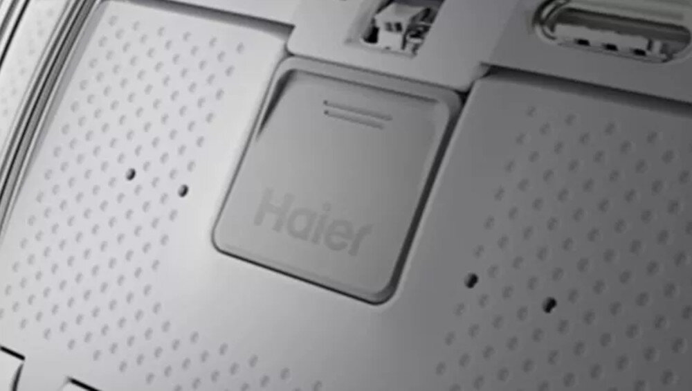 Zbliżenie na panel pralki z widocznym logo Haier. Panel ma perforowaną powierzchnię i prostokątny element z napisem Haier.