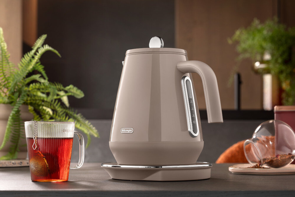 Na blacie kuchennym stoi beżowy czajnik elektryczny z logo \'DeLonghi\', obok znajduje się szklanka z herbatą. W tle widoczne są zielone rośliny i elementy kuchenne.