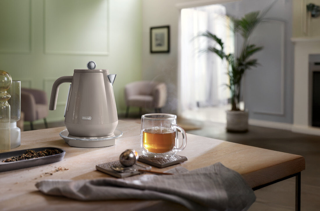 Na drewnianym stole stoi beżowy czajnik elektryczny z logo \'DeLonghi\', obok szklanka z herbatą na podstawce. W tle widać rozmyty salon z rośliną doniczkową i fotelami.