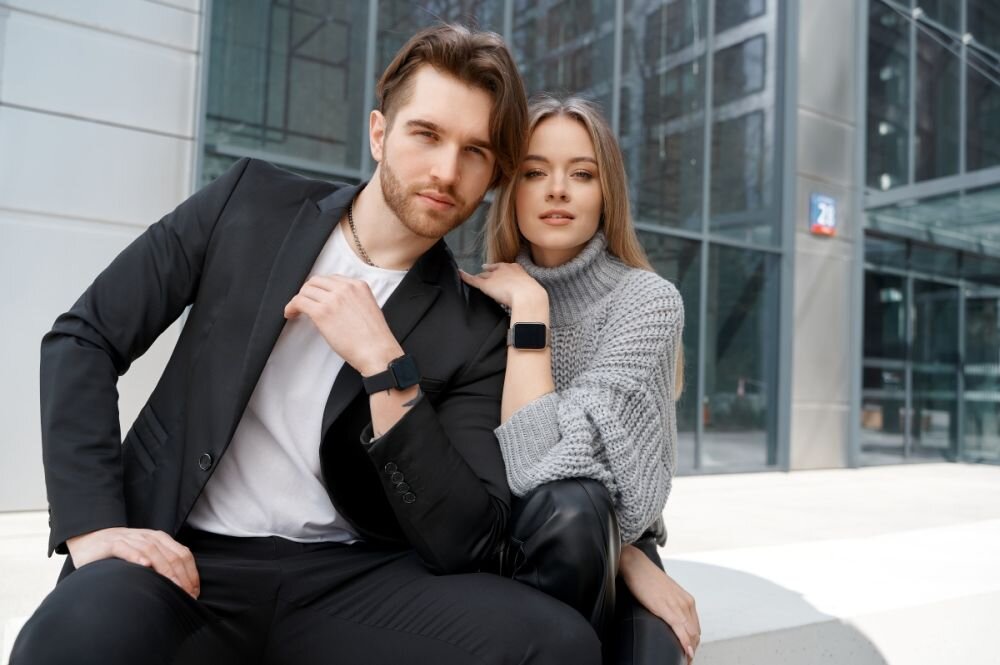 Dwie osoby siedzą na zewnątrz przed nowoczesnym budynkiem, obie noszą czarne smartwatche na nadgarstkach. Osoby są ubrane elegancko, jedna w czarną marynarkę, druga w szary sweter.