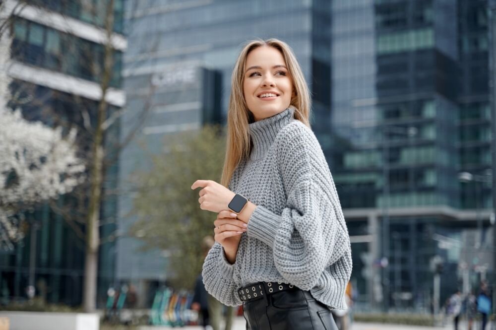 Osoba w szarym swetrze stoi na tle nowoczesnych budynków, nosząc czarny smartwatch na nadgarstku. W tle widoczne są drzewa i szklane fasady budynków.