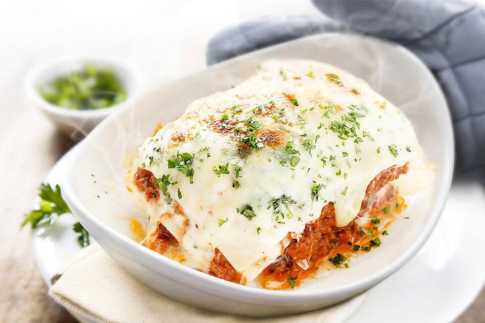 Parująca lasagne widoczna na zdjęciu ilustruje funkcję podtrzymywania ciepła gotowych potraw Na białym talerzu znajduje się porcja lasagne z widocznym roztopionym serem i posypką z ziół. W tle widoczna jest miseczka z zielonymi ziołami oraz rękawica kuchenna.