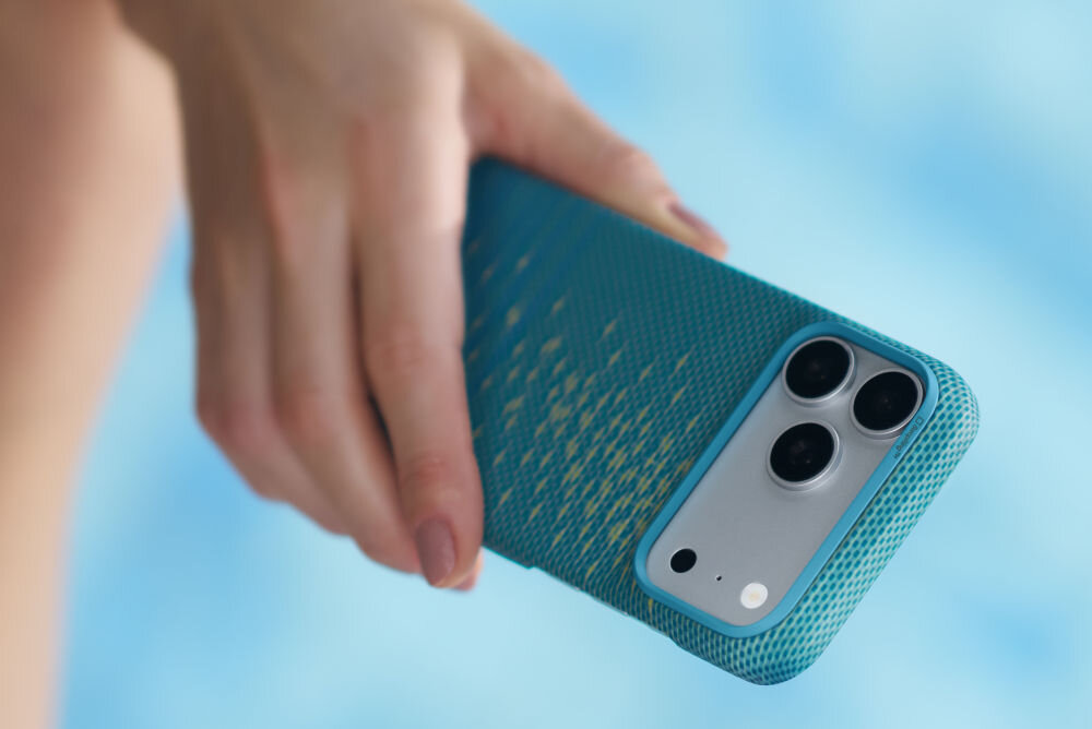 Osoba trzyma telefon w niebieskim etui z teksturą, widoczne są trzy obiektywy aparatu i lampa błyskowa. Tło jest rozmyte i ma jasnoniebieski kolor.