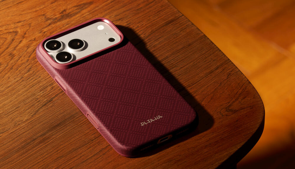 Na drewnianym stole leży telefon w burgundowym etui z trzema widocznymi obiektywami aparatu. Na etui widnieje logo \'PITAKA\'.