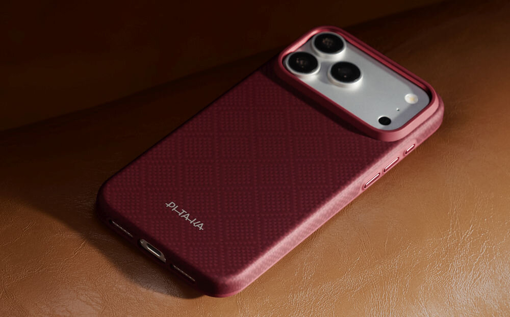 Burgundowe etui na telefon z trzema widocznymi obiektywami aparatu i logo \'PITAKA\' na dole. Etui leży na brązowej powierzchni.