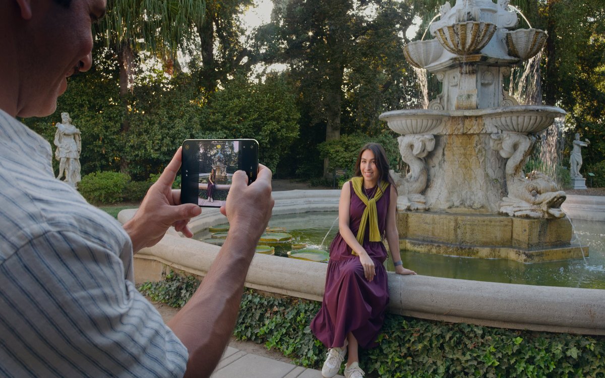 Osoba fotografująca kobietę siedzącą blisko fontanny używając iPhone Duo.