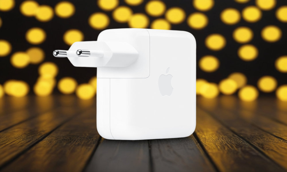 Biała ładowarka sieciowa z logo Apple, umieszczona na drewnianej powierzchni z rozmytym tłem w żółte kropki.