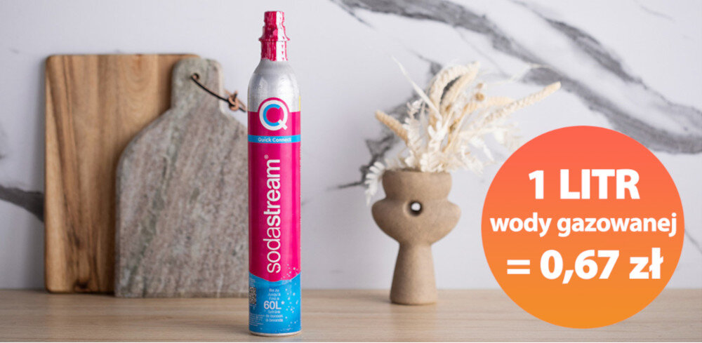 Na blacie stoi butla z napisem \'sodastream\', obok której znajduje się okrągła grafika z tekstem \'1 LITR wody gazowanej = 0,67 zł\'. W tle widoczne są deski do krojenia i wazon z suszonymi roślinami.