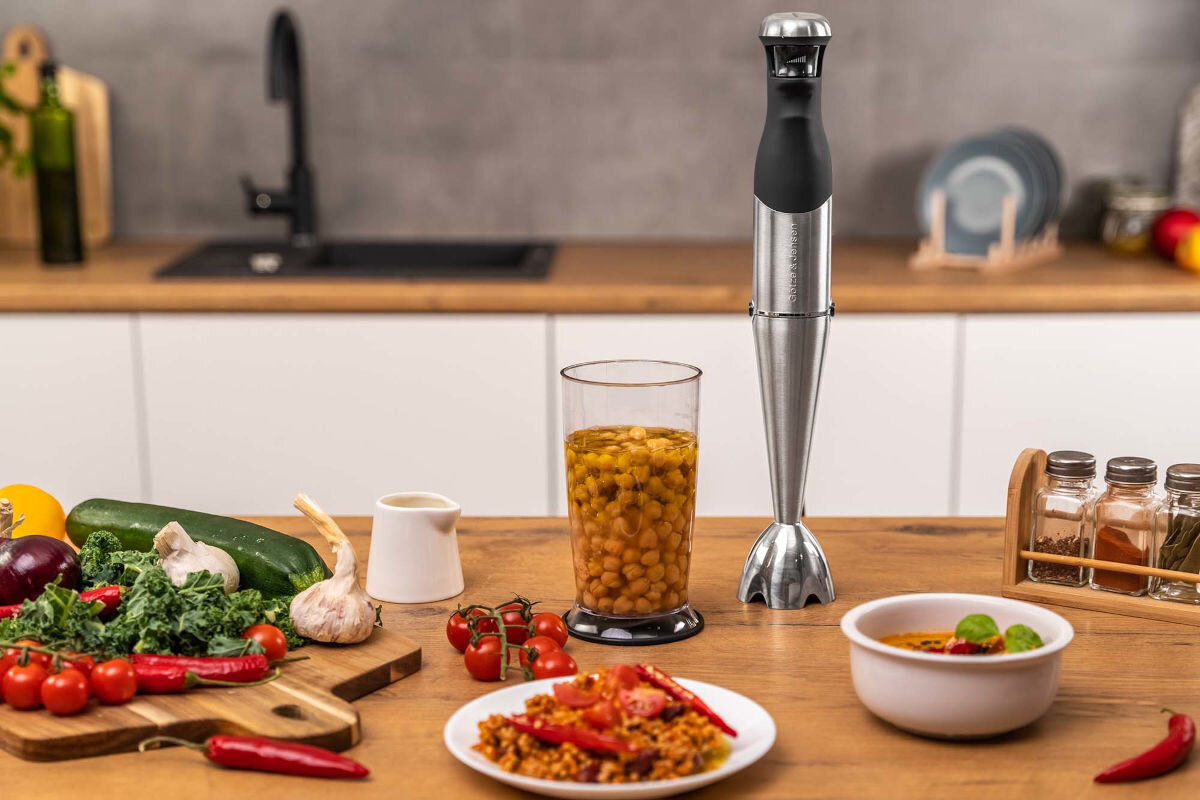 Na blacie kuchennym stoi blender ręczny w kolorze inox, obok niego pojemnik z ciecierzycą. Wokół znajdują się różne warzywa, przyprawy i miska z potrawą.