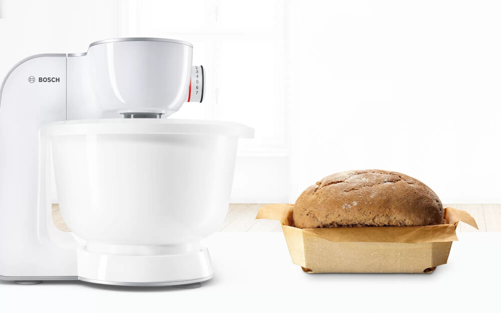 Na obrazie widoczny jest biały robot kuchenny z misą oraz bochenek chleba w papierowej foremce. Na urządzeniu znajduje się logo \'BOSCH\'.