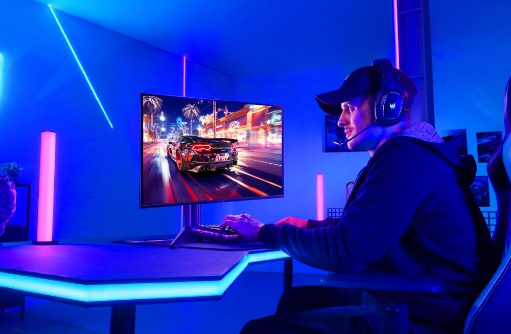 Osoba siedzi przy biurku, grając na komputerze z dużym monitorem, na którym wyświetlana jest dynamiczna scena z gry wyścigowej. Pomieszczenie jest oświetlone niebieskimi i różowymi światłami LED.