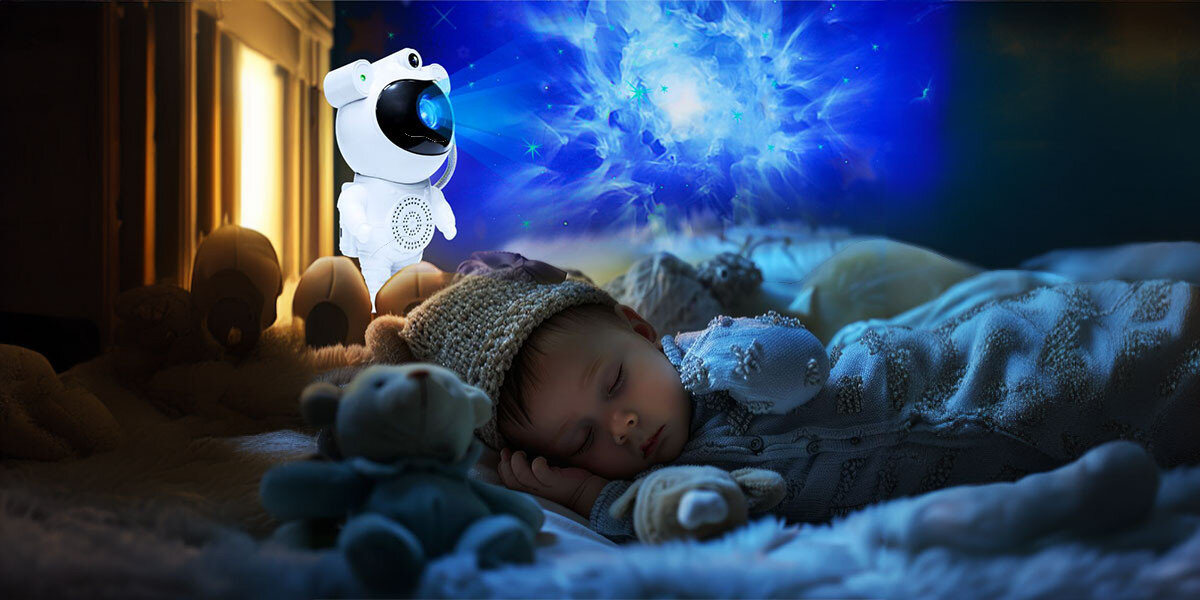 Na obrazie widać dziecko śpiące w łóżku, obok którego znajduje się projektor w kształcie astronauty. Na ścianie widoczna jest projekcja gwiazd w niebieskich odcieniach.