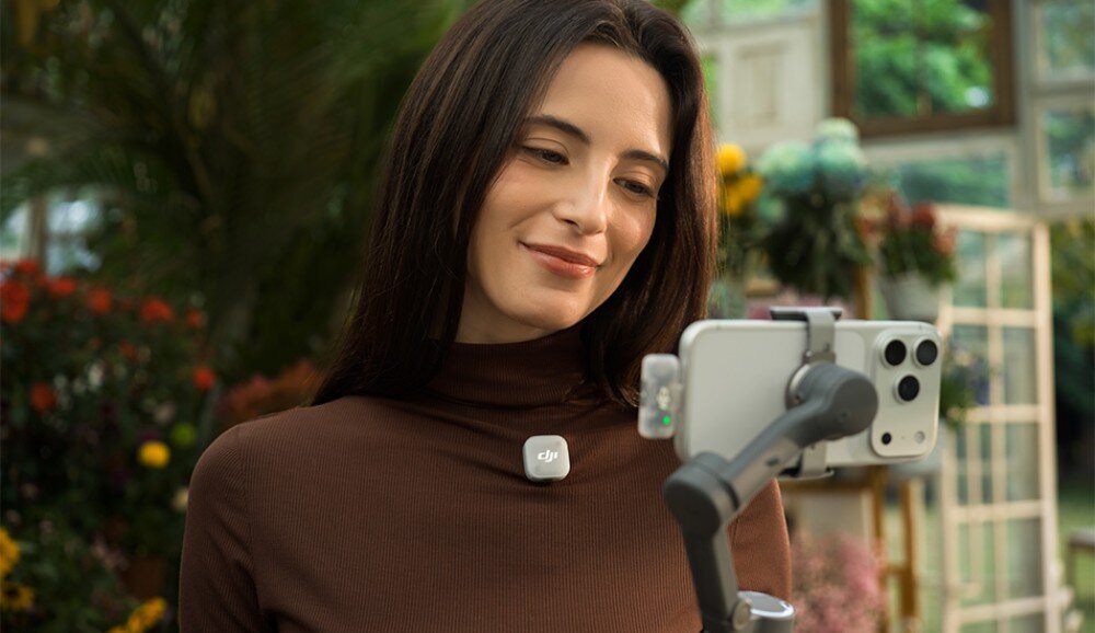 Osoba trzymająca smartfon na stabilizatorze, z przypiętym mikrofonem z logo \'dji\' na ubraniu. W tle widoczne są rośliny i kwiaty.