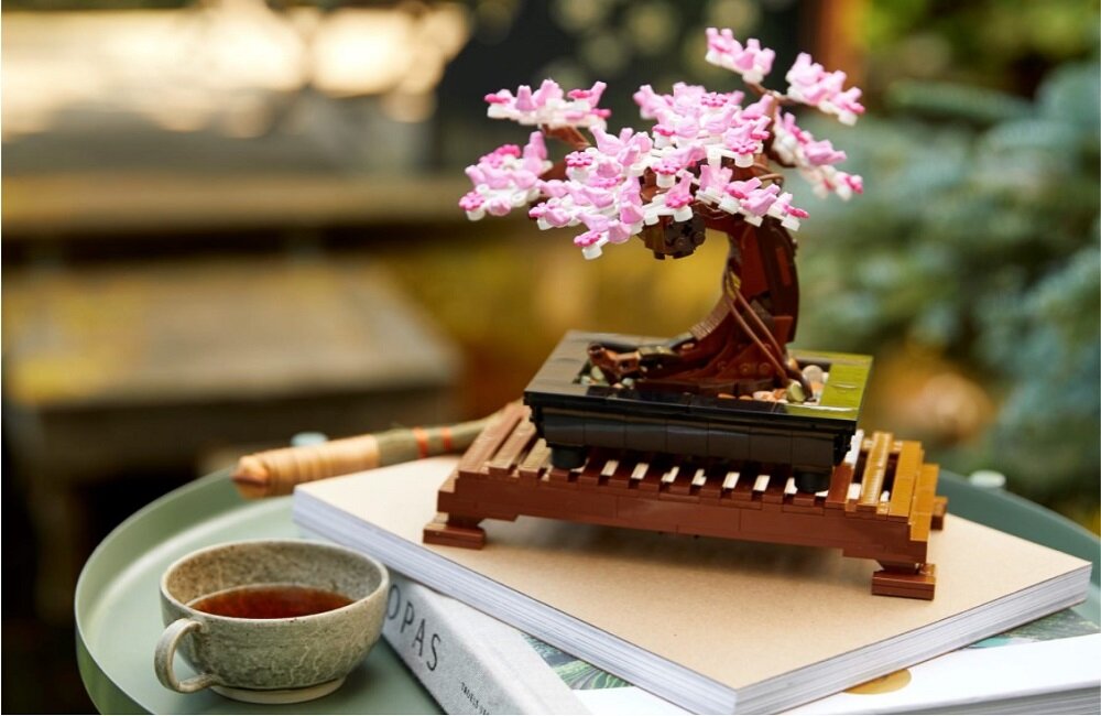 Model drzewka bonsai z różowymi kwiatami stoi na drewnianej podstawce. Obok znajduje się filiżanka z herbatą i książka z napisem \'OPAS\'.