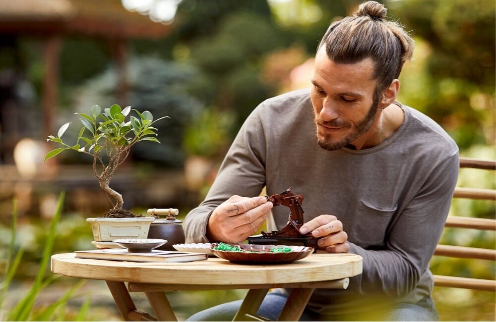 Na okrągłym drewnianym stole znajduje się małe drzewko bonsai w doniczce oraz kilka talerzyków z elementami. W tle widoczny jest rozmyty ogród.