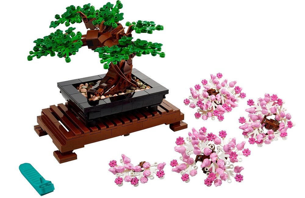 Zestaw LEGO przedstawiający drzewko bonsai z zielonymi liśćmi na czarnej podstawce umieszczonej na brązowej platformie. Obok znajdują się różowe elementy kwiatowe i zielone narzędzie.