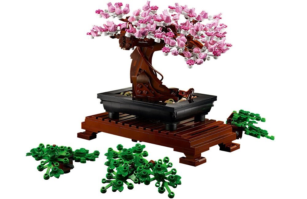 Model drzewka bonsai z różowymi kwiatami w czarnej doniczce na brązowej podstawce. Obok znajdują się zielone elementy przypominające liście.