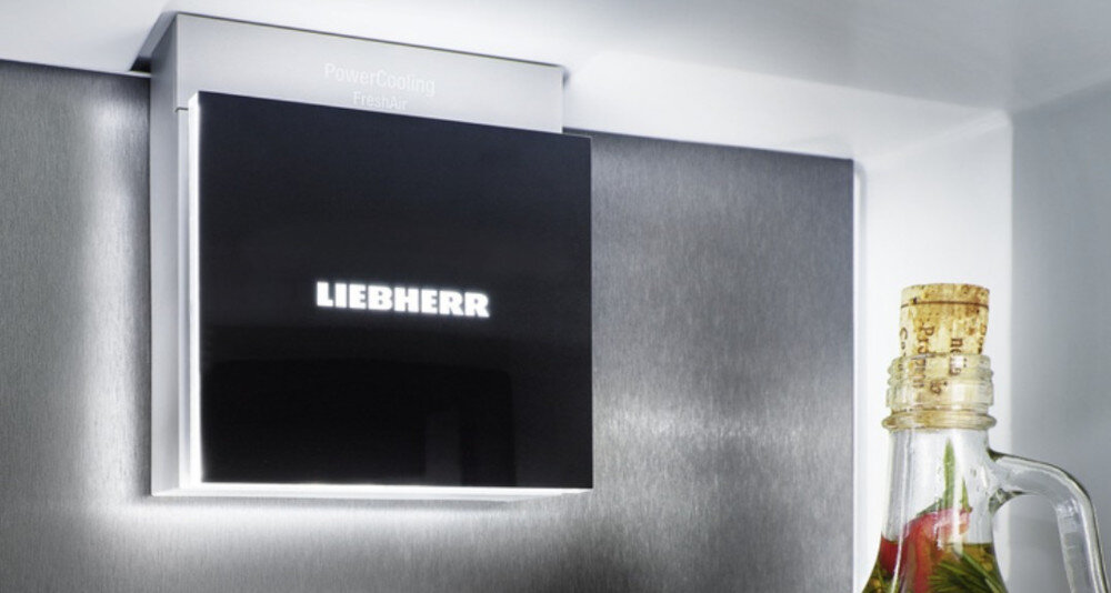 Na obrazie widoczny jest fragment lodówki z logo LIEBHERR na czarnym panelu. Obok znajduje się butelka z korkiem i kolorowym płynem w środku.