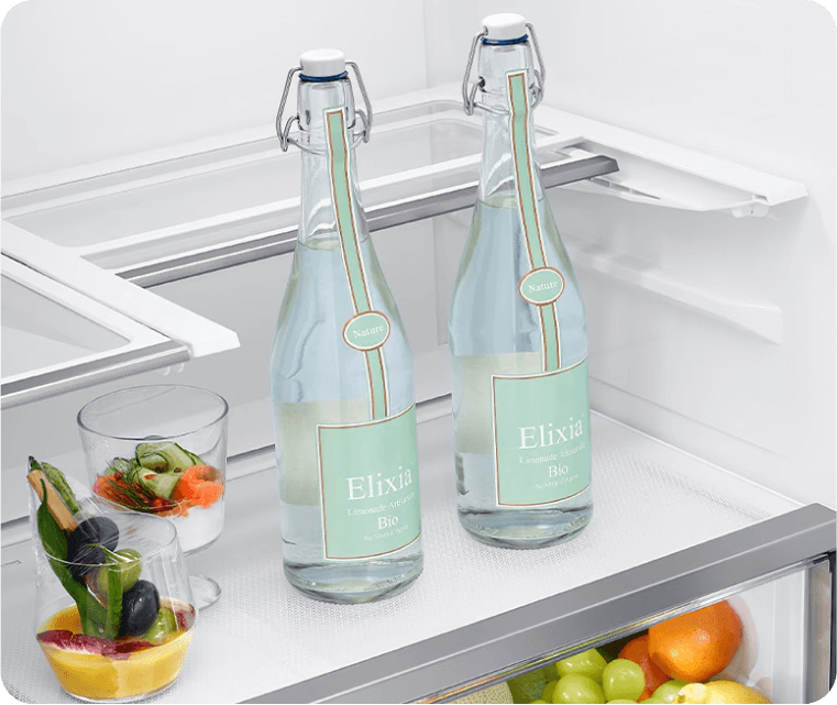 Wnętrze lodówki z dwiema wysokimi butelkami wody. W lodówce znajdują się dwie butelki z etykietą \'Elixia Limonade Artisanale Bio\', szklanka z warzywami oraz pojemnik z owocami. Wnętrze lodówki jest jasne i przestronne.