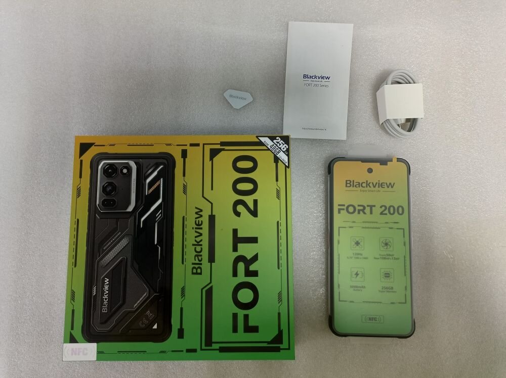 Na obrazie znajduje się pudełko z napisem \'Blackview FORT 200\' oraz smartfon z wyświetlaczem pokazującym ten sam napis. Obok leży kabel, instrukcja obsługi i mały element z napisem \'Blackview\'.