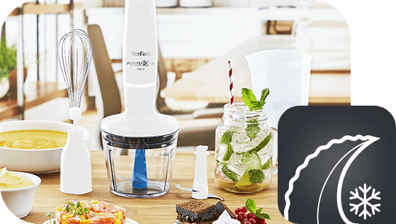 Na drewnianym stole znajduje się blender ręczny z logo Tefal, obok którego leży trzepaczka i różne potrawy. W prawym dolnym rogu widoczna jest ikona z symbolem liścia i płatka śniegu.