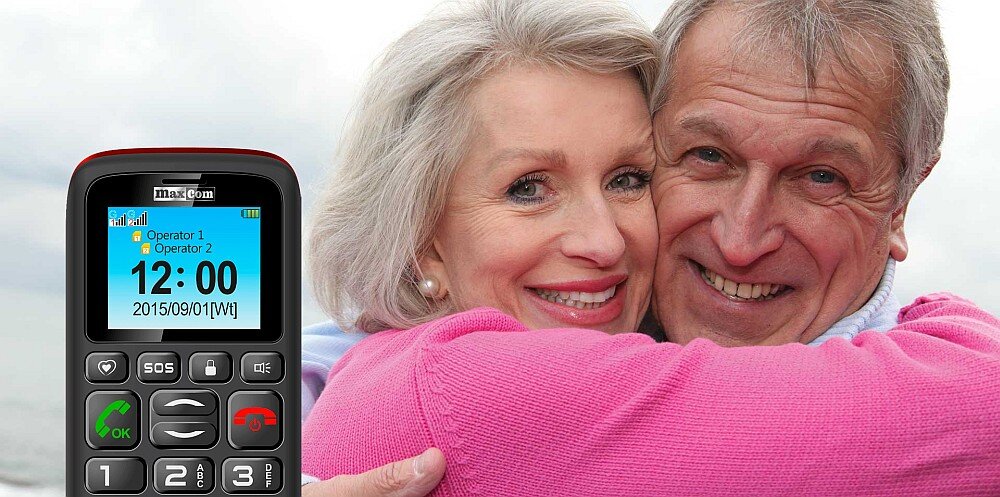 Na obrazie widać telefon z dużymi przyciskami i wyświetlaczem pokazującym godzinę 12:00 oraz datę 2015/09/01 [Wt]. Obok telefonu znajduje się para starszych osób obejmujących się i uśmiechających.