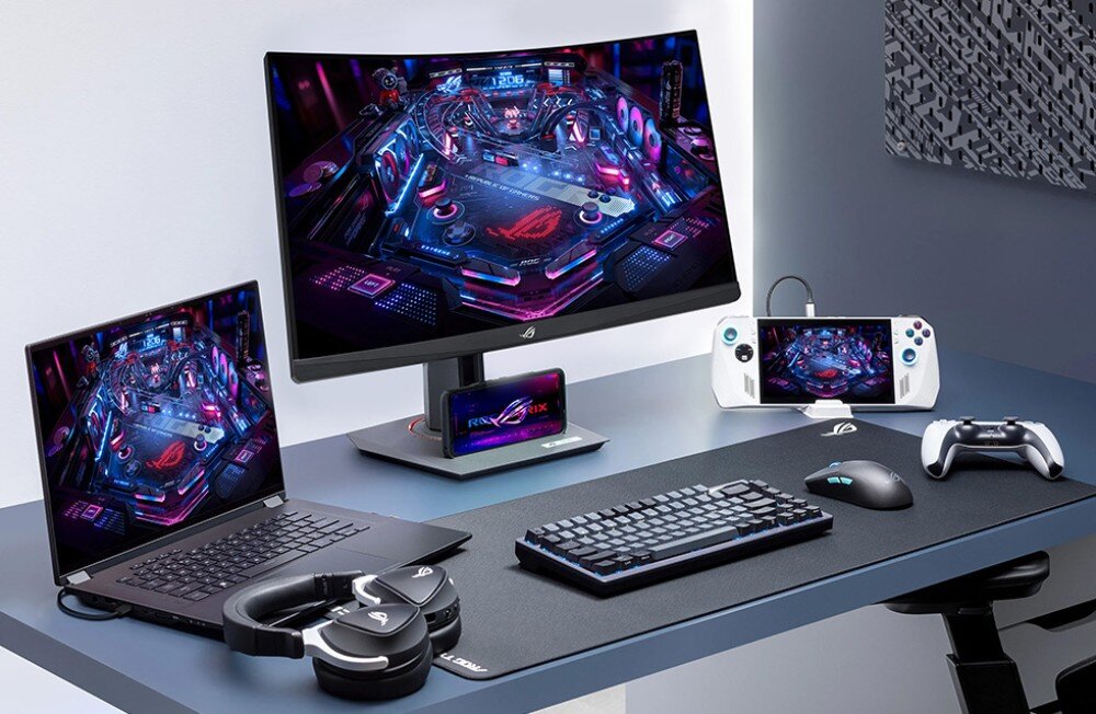 Na biurku znajduje się monitor z wyświetlonym kolorowym obrazem, obok laptop, klawiatura, mysz, słuchawki, kontroler i urządzenie przenośne. Na monitorze i laptopie widoczne jest logo producenta w formie czerwonego symbolu na czarnym tle.
