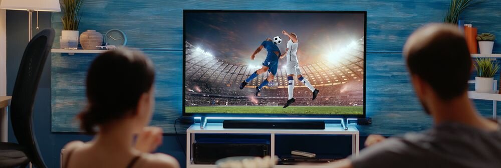 Dwie osoby siedzą przed telewizorem, na którym wyświetlany jest mecz piłki nożnej. Pod telewizorem znajduje się czarny soundbar.