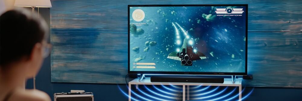 Osoba ogląda telewizor, na którym wyświetlana jest gra z motywem kosmicznym. Pod telewizorem znajduje się czarny soundbar, a wokół niego widoczne są efekty dźwiękowe w postaci fal.