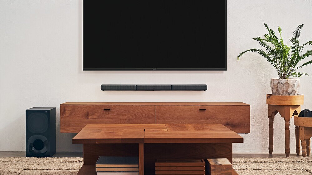 W salonie znajduje się duży telewizor zamontowany na ścianie, poniżej którego umieszczony jest czarny soundbar. Obok drewnianej szafki stoi subwoofer, a na podłodze leży dywan.