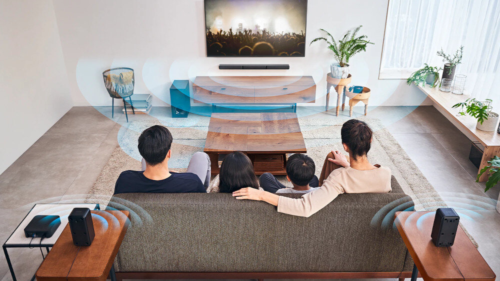 W salonie znajduje się telewizor z zamontowanym soundbarem na ścianie, przed którym siedzi grupa osób na kanapie. Wokół rozstawione są głośniki, a na ekranie telewizora wyświetlany jest koncert.