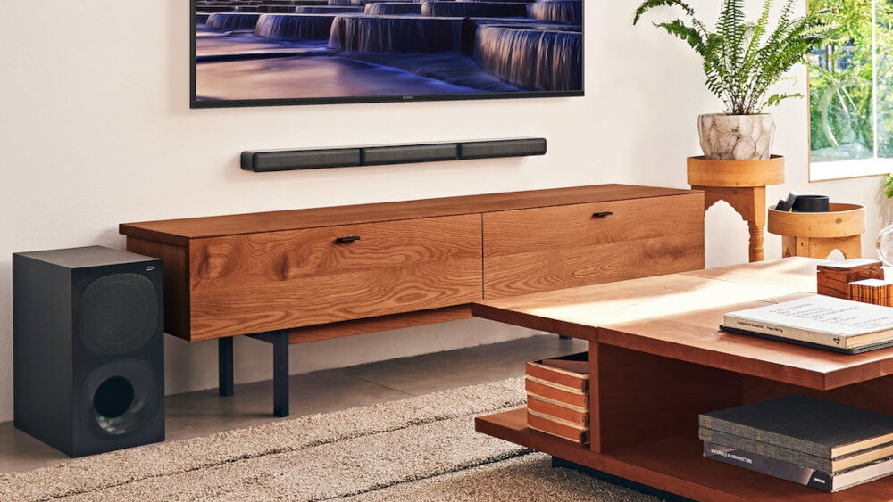 W salonie znajduje się drewniana szafka z telewizorem na ścianie oraz soundbar i subwoofer. Obok stoi stolik z książkami i rośliną w doniczce.