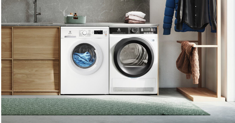 Na obrazie znajduje się pralka i suszarka marki Electrolux ustawione obok siebie w nowoczesnej pralni. Pralka ma otwarte drzwi z widocznym niebieskim ręcznikiem w środku, a suszarka ma przezroczyste drzwi z widocznym bębnem.