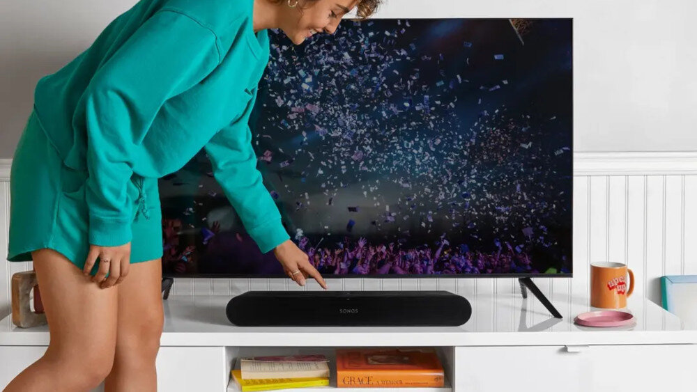 Osoba w zielonym stroju dotyka czarnego soundbara marki SONOS umieszczonego pod telewizorem, na którym wyświetlany jest obraz z koncertu. Na półce obok soundbara znajdują się książki i kubek.