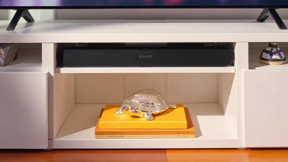 Czarny soundbar z logo \'SONOS\' umieszczony na półce białego mebla. Pod soundbarem znajduje się przezroczysta figurka żółwia na pomarańczowej książce.