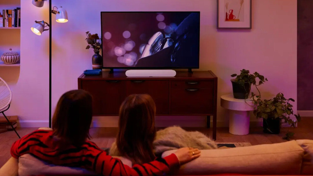 Dwie osoby siedzą na kanapie przed telewizorem, na którym wyświetlany jest film. Przed telewizorem znajduje się czarny soundbar na drewnianej szafce, a w pomieszczeniu są rośliny i lampa stojąca.