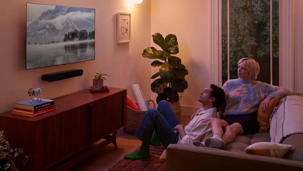 W salonie na ścianie wisi telewizor z wyświetlonym krajobrazem górskim, a pod nim zamontowany jest czarny soundbar. Na kanapie siedzą dwie osoby, a obok stoi roślina doniczkowa i szafka z książkami oraz dekoracją.