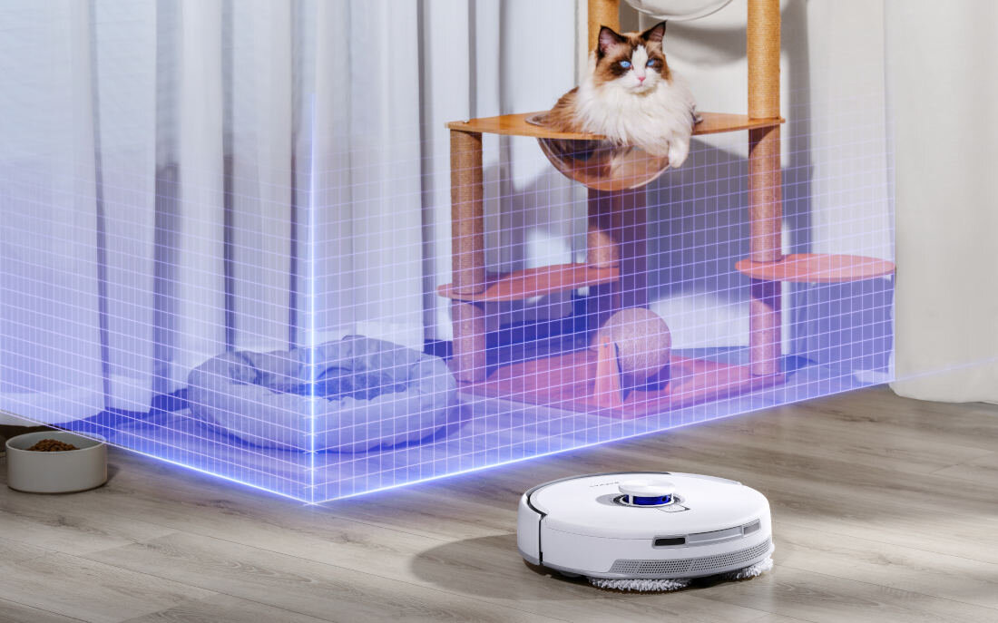 Na podłodze znajduje się biały robot sprzątający, a w tle widać kota siedzącego na drapaku. Obok drapaka znajduje się legowisko dla zwierząt.