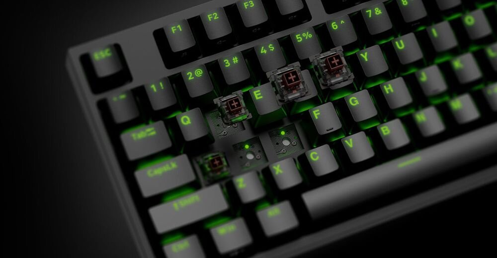 Czarna klawiatura mechaniczna z podświetleniem RGB, widoczne są klawisze z oznaczeniami liter i cyfr oraz kilka zdjętych nakładek klawiszy, ukazujących mechaniczne przełączniki. Na klawiszach widoczne są oznaczenia takie jak \'Esc\', \'Q\', \'E\', \'Z\', \'X\', \'C\'.