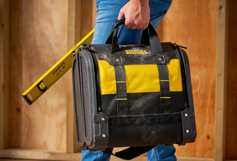 Torba narzędziowa STANLEY FatMax 1-94-231 pomieszczenie drewniana ściana osoba niesie torbę w dłoni w drugiej dłoni trzyma poziomicę przechowywanie i transport narzędzi wszystkie akcesoria pozostają w jednym miejscu porządek solidna konstrukcja funkcjonalne rozwiązania