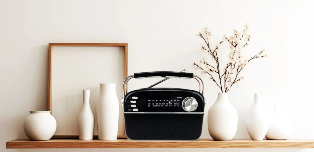 Na drewnianej półce stoi czarne, przenośne radio z analogowym tunerem i dużym pokrętłem. Obok radia znajdują się białe wazony i gałązka z kwiatami.