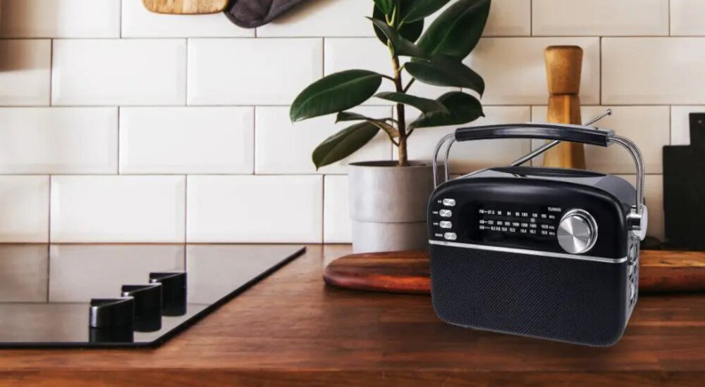 Na drewnianym blacie kuchennym stoi czarne, przenośne radio z widocznym panelem sterowania i anteną. W tle widać białe kafelki, roślinę w doniczce oraz elementy kuchenne.