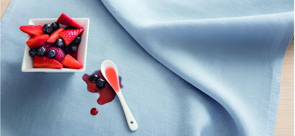 Na jasnoniebieskim obrusie znajduje się biała miseczka z owocami, takimi jak truskawki i jagody. Obok leży biała łyżeczka z czerwonym sosem oraz kilka kropli sosu na obrusie.